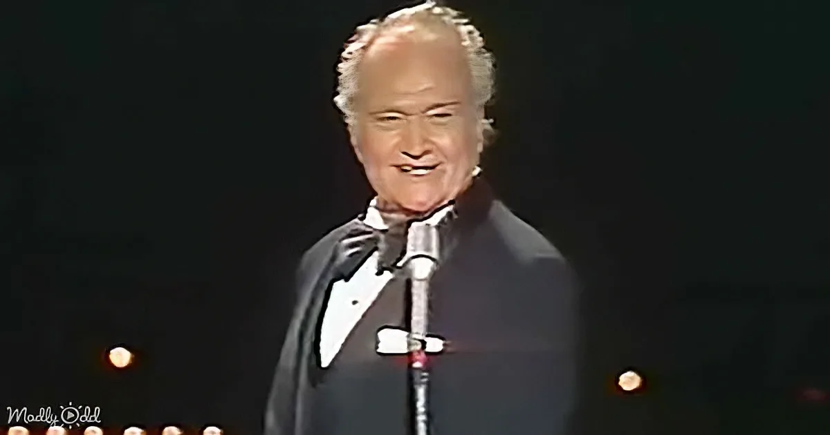 A man in a tuxedo smiles on stage, holding a microphone, with a spotlight and dark background.