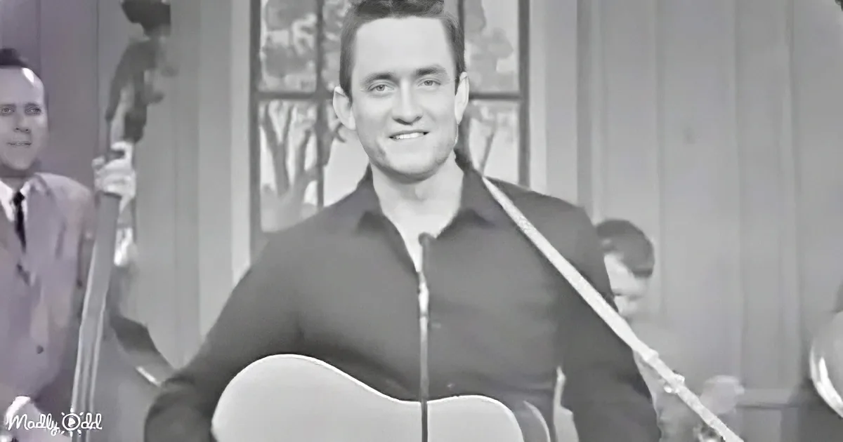 A man in a black shirt smiles while holding an acoustic guitar, with a band in the background.