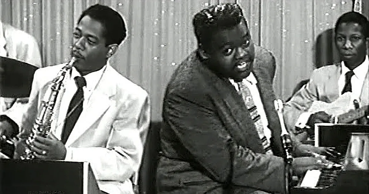 A man in a suit plays a saxophone while another man in a patterned suit sits nearby, possibly playing a keyboard.