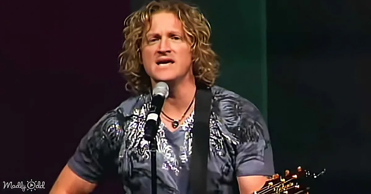 A man with curly hair sings into a microphone while playing a guitar on stage.
