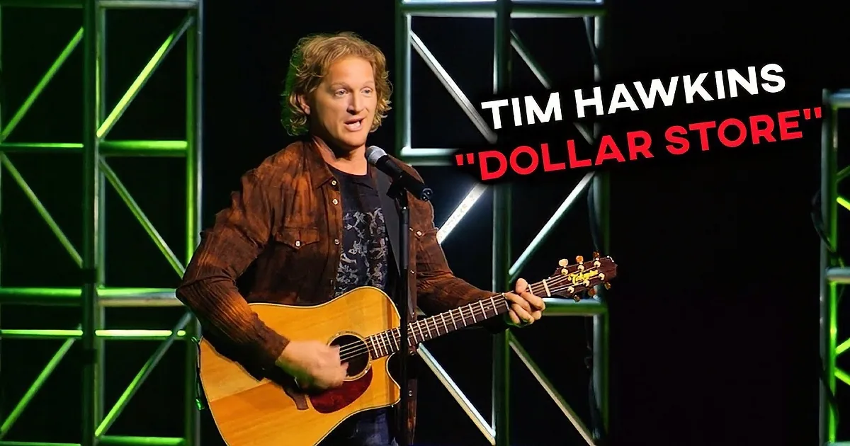 A man in a brown jacket plays an acoustic guitar on stage with green lighting.