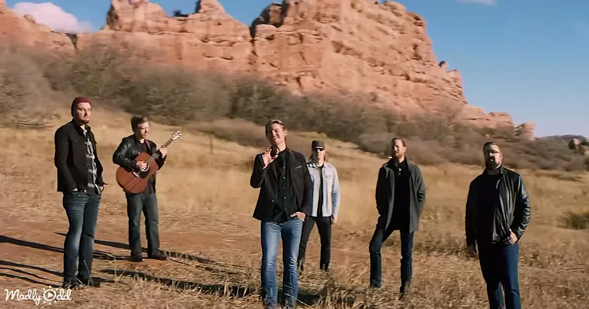 Six men stand in a field with a guitarist and singer, against a backdrop of red rock formations.