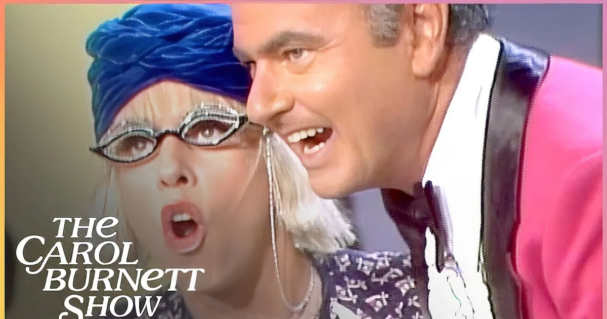 A man in a pink suit leans close to a woman wearing a blue headscarf and large glasses, both appearing surprised.