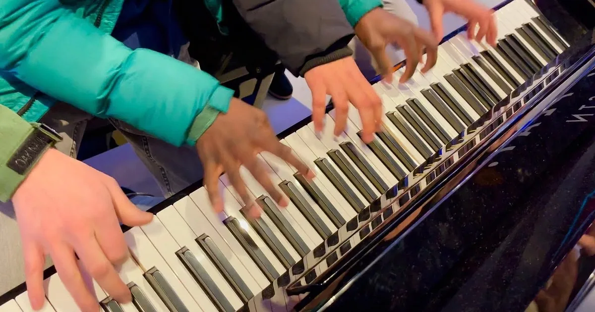 Three people play a piano together, surrounded by onlookers in a public space.