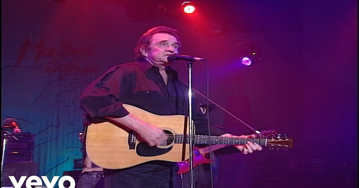 A man sings into a microphone while playing an acoustic guitar on stage under red and purple lights.