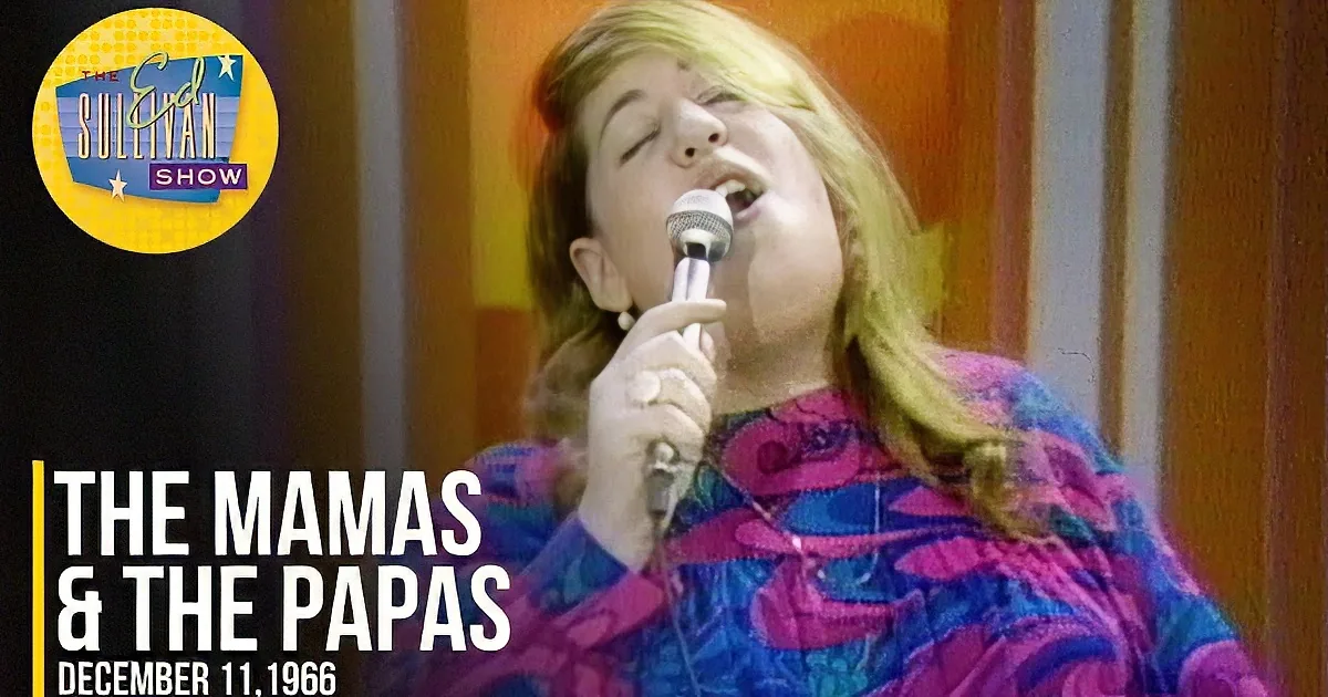 A woman with long blonde hair speaks into a microphone, wearing a colorful patterned top, against a vibrant background.