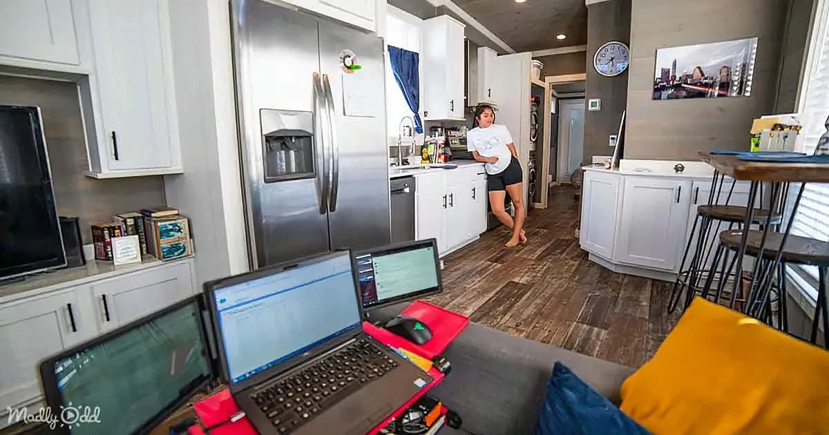 A woman stands in a modern kitchen, surrounded by laptops and a cozy living space with a yellow pillow.