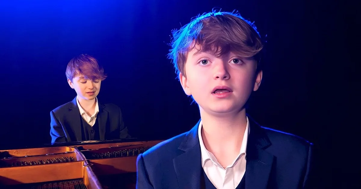 A young boy in a suit plays a grand piano under stage lighting.