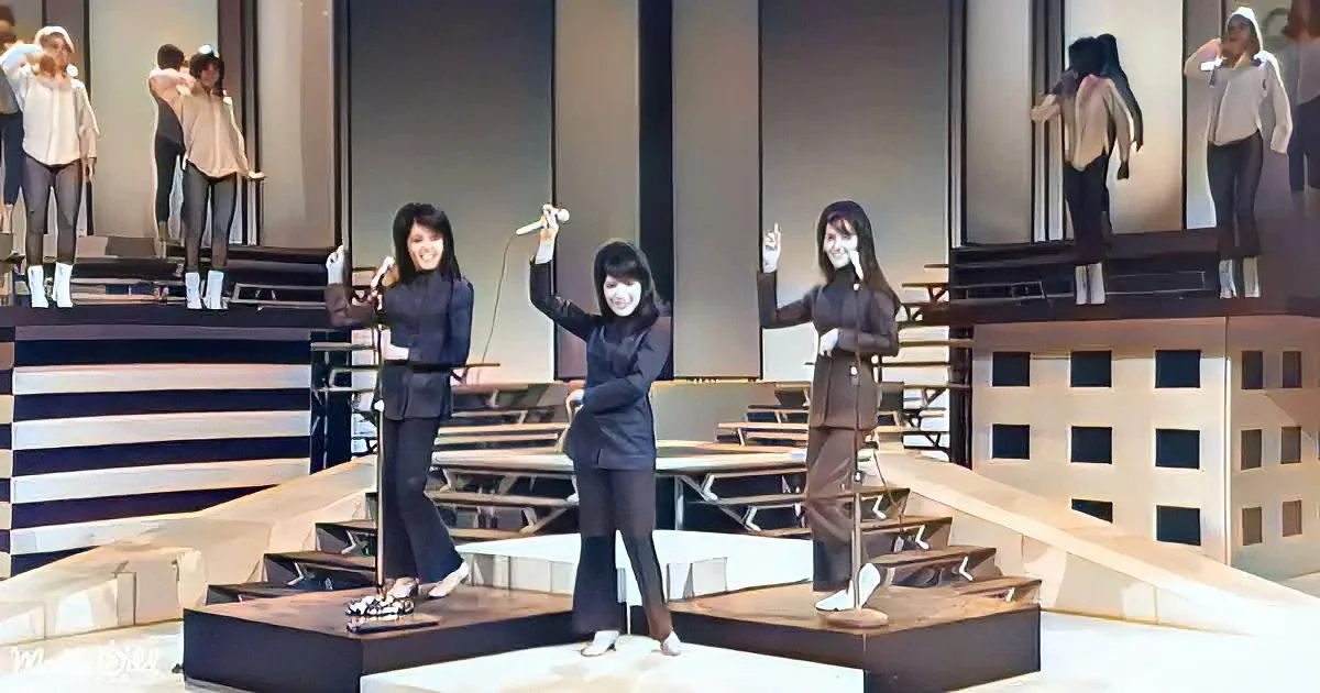 Three women in black outfits perform on stage with microphones, surrounded by large windows and cityscape.