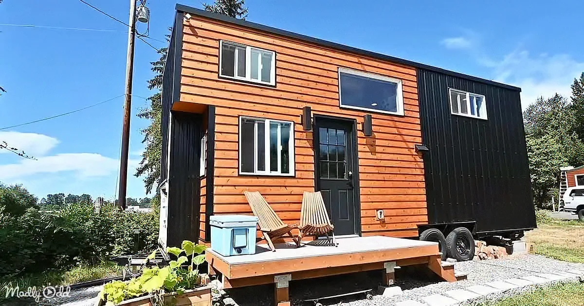 A modern tiny house with orange and black siding, a wooden deck, and two chairs, set against a backdrop of trees and a...