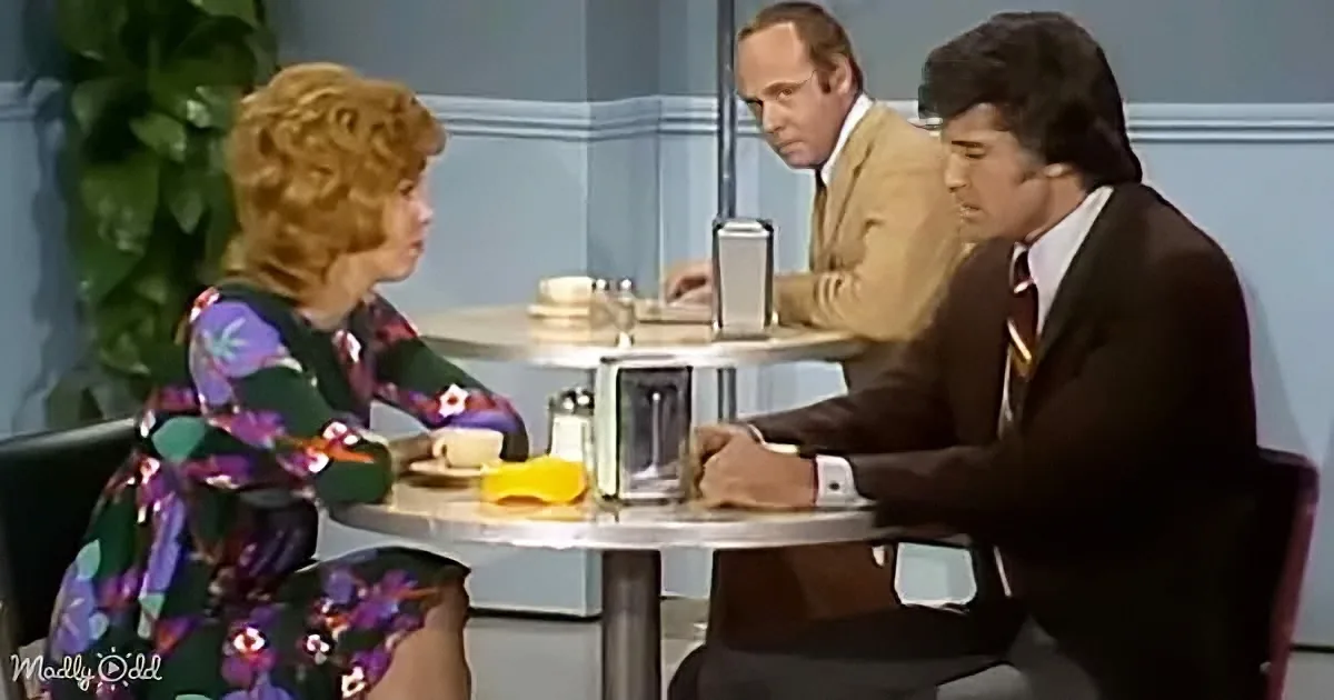 A woman in a floral dress sits at a table with a man in a suit, while another man in a tan jacket looks on.