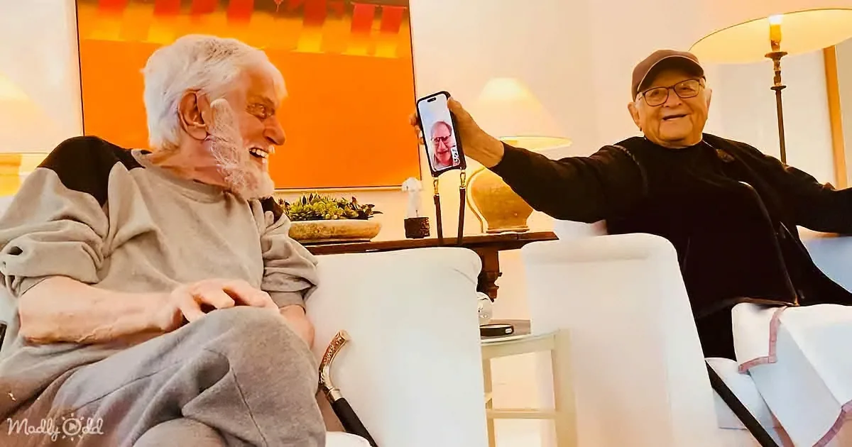 Two elderly men are sitting on a couch, one holding a phone showing a video call with another man.