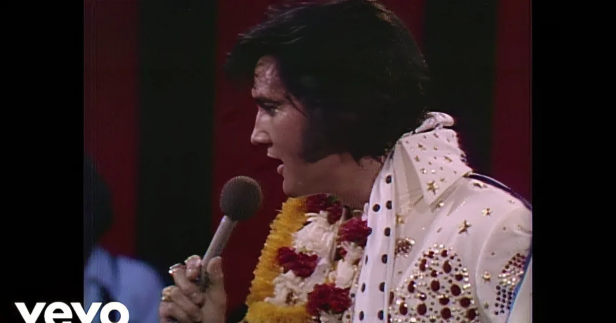A performer in a white sequined jacket and red scarf sings into a microphone.
