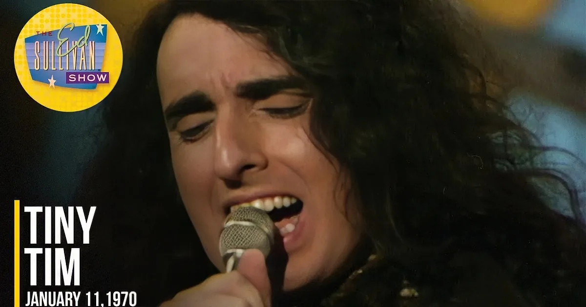 A person with long, dark, curly hair sings into a microphone, eyes closed, against a blurred, colorful background.