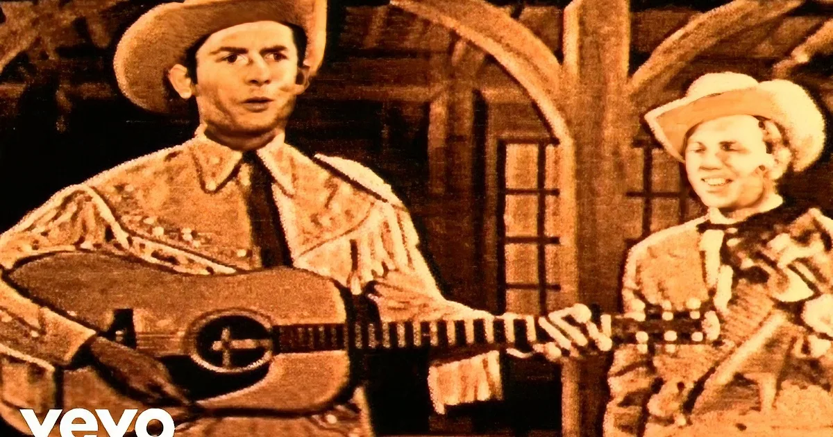 A man in a cowboy hat plays guitar while another in a similar outfit sings nearby.