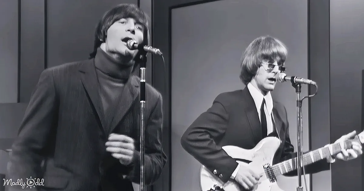 A man in a suit sings into a microphone, another plays guitar, both in a black-and-white setting.