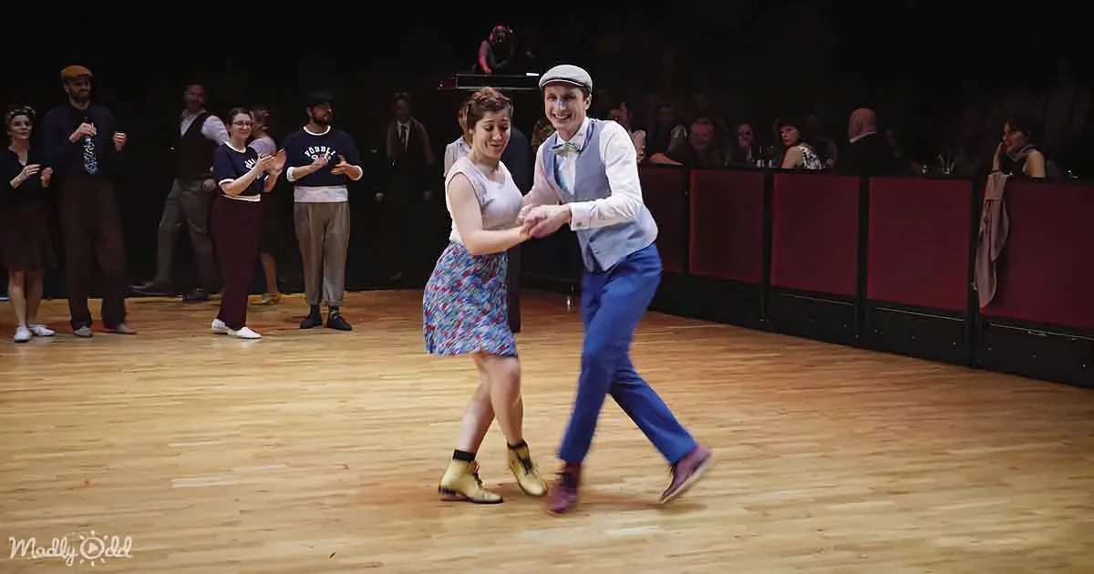 A couple in vintage attire dances joyfully on a wooden floor, surrounded by onlookers.