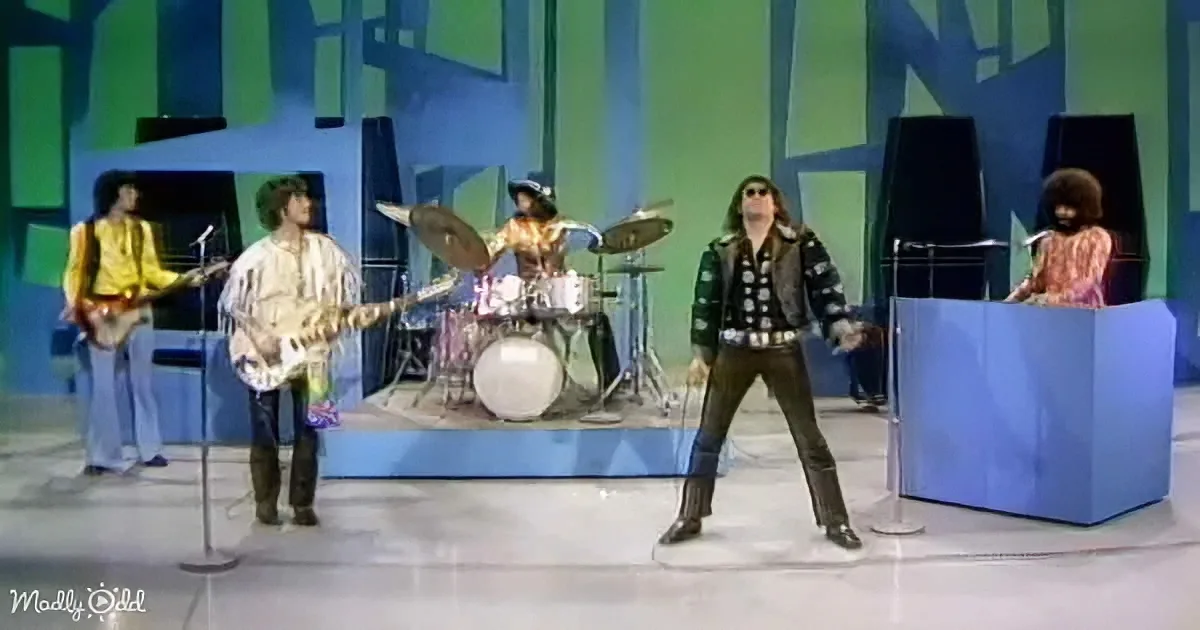 A band performs on a stage with colorful background, featuring a drummer, guitarist, keyboardist, and lead singer.