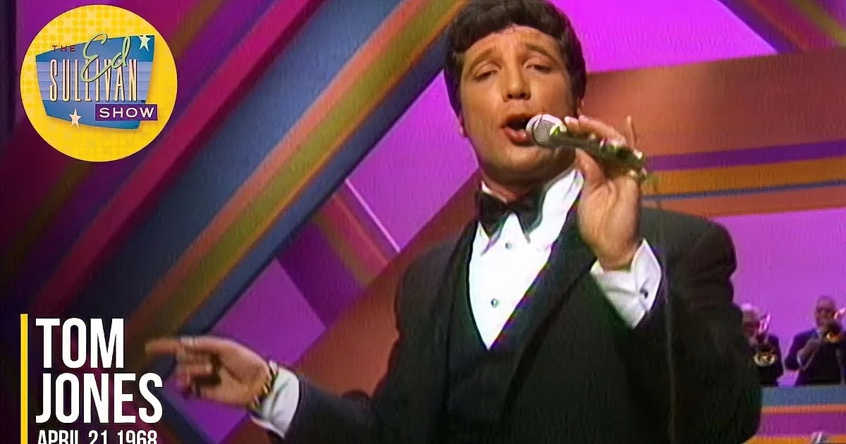A man in a tuxedo sings into a microphone on stage with a colorful background and musicians.
