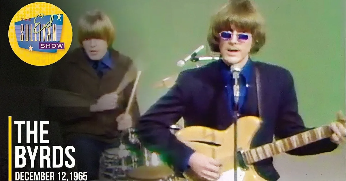 A man in a suit plays guitar while another in a brown jacket plays drums.