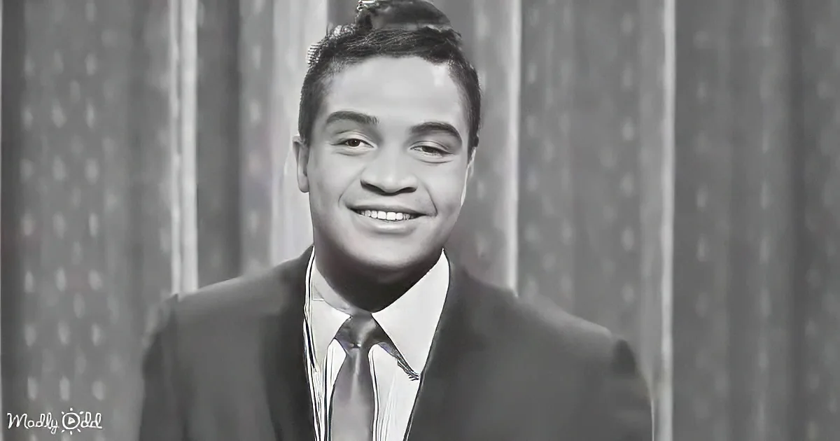 A smiling man in a suit and tie, with a neatly combed hairstyle, stands against a patterned backdrop.