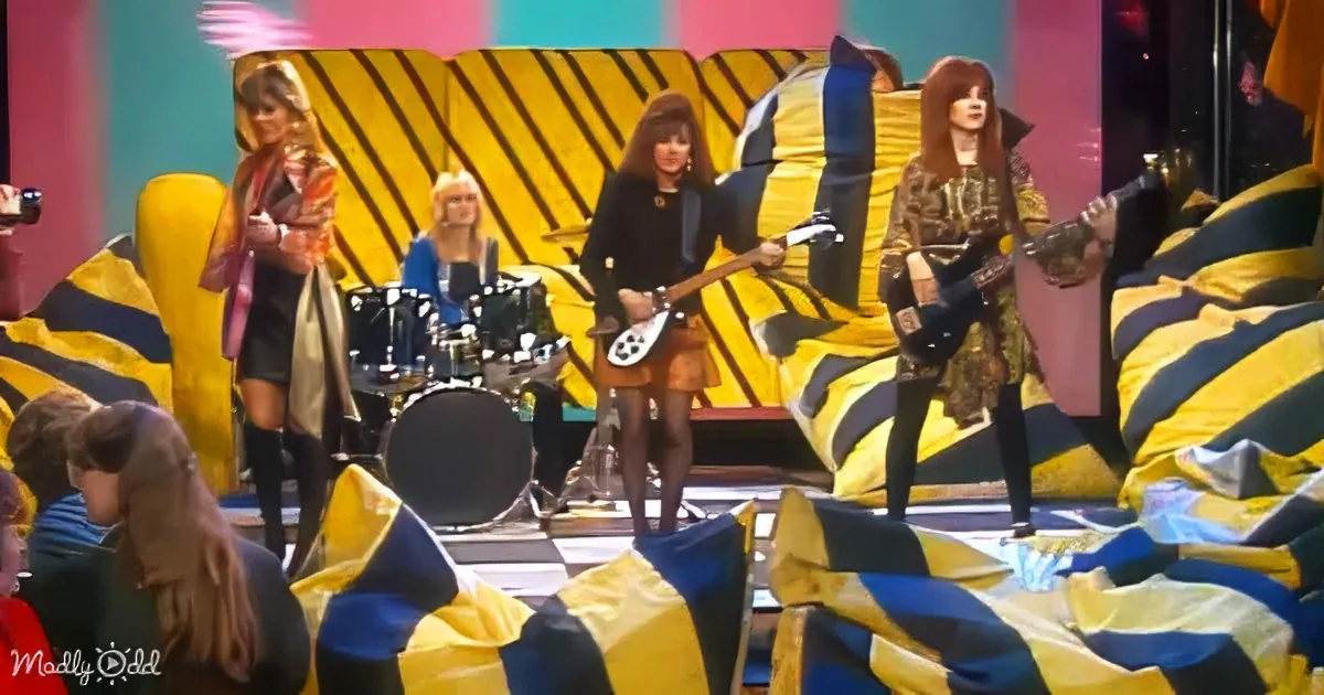 A band performs on stage with a colorful, striped backdrop and audience in the foreground.
