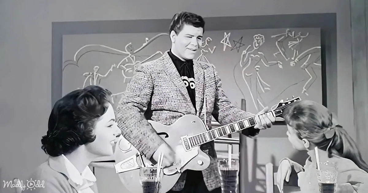 A man in a textured suit plays a guitar in front of two women, with a chalkboard behind him.