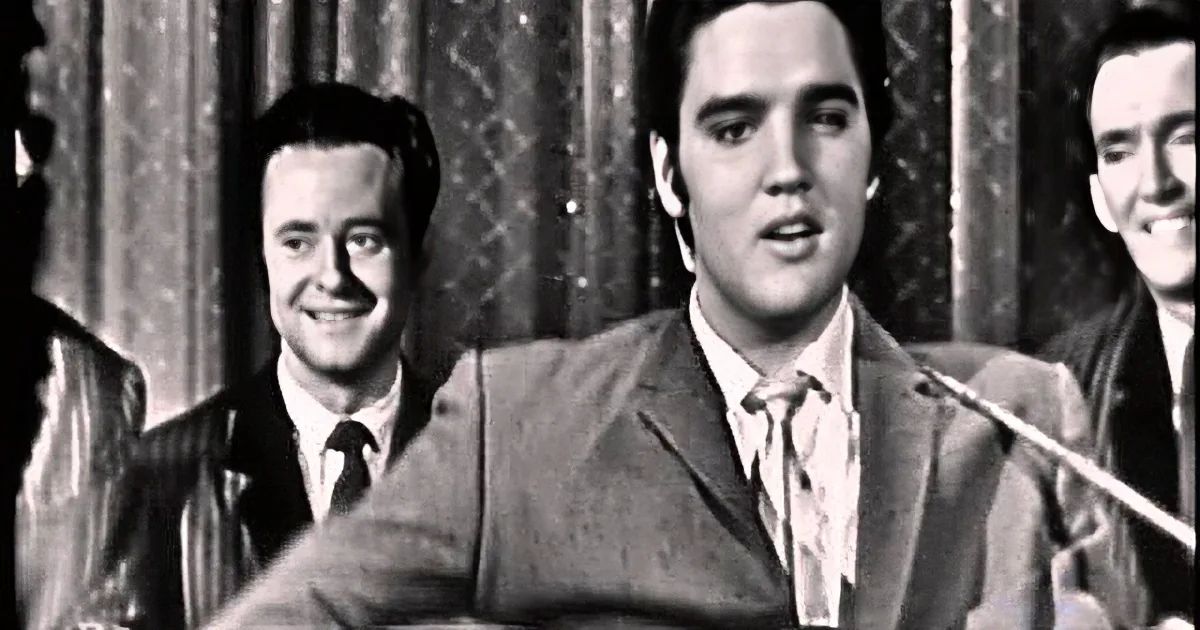 Elvis Presley in a black-and-white scene, surrounded by two men, with a curtain backdrop.