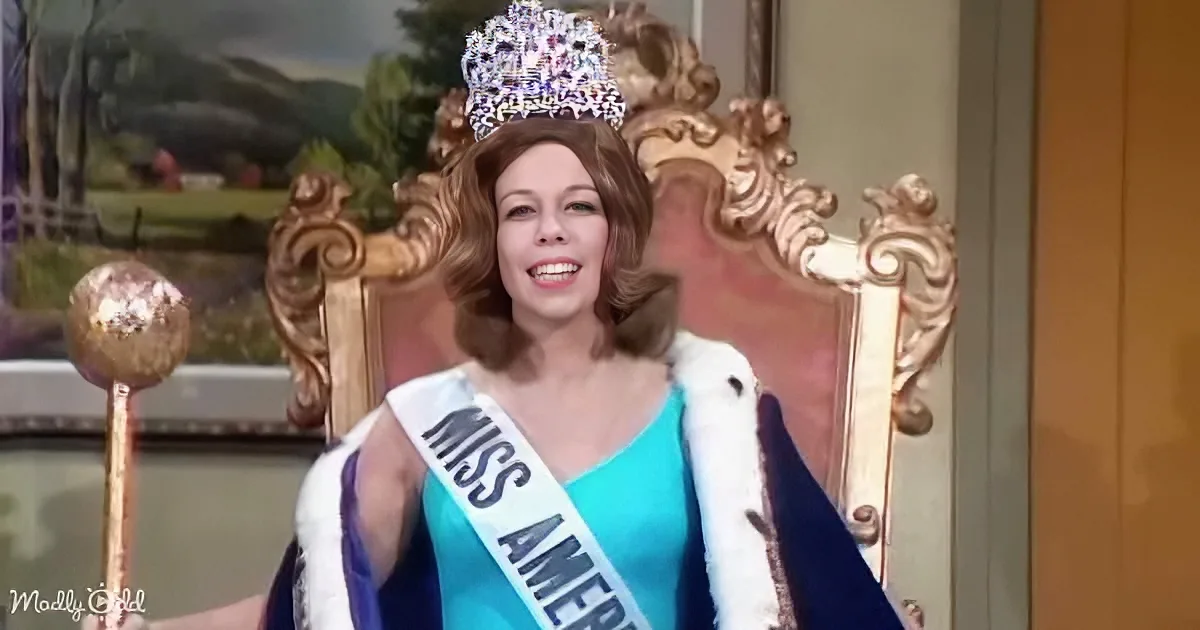A woman in a blue dress and sash, wearing a crown, sits on a throne-like chair, holding a scepter.