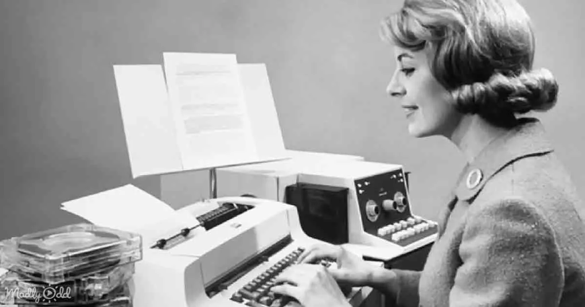 A woman types on a vintage computer, surrounded by papers and a tape recorder.