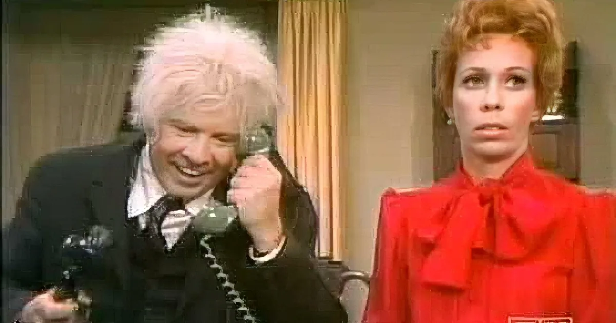 A man in a suit and white hair is laughing while holding a phone, next to a woman in a red dress.