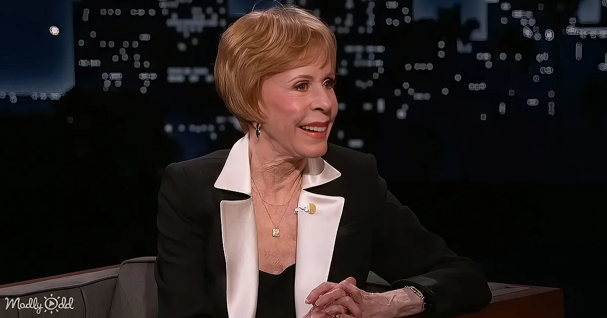 A woman with short blonde hair, wearing a black blazer and white shirt, smiles while sitting on a couch.