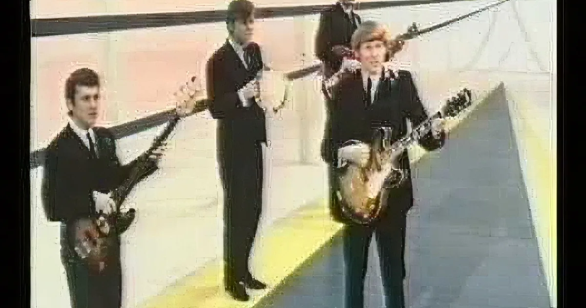 A man in a black suit and tie plays a guitar, singing passionately, with two other musicians in the background.
