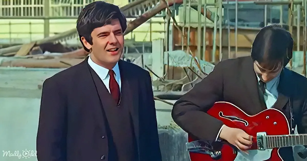 A man in a suit sings while another plays a red guitar in a construction site backdrop.