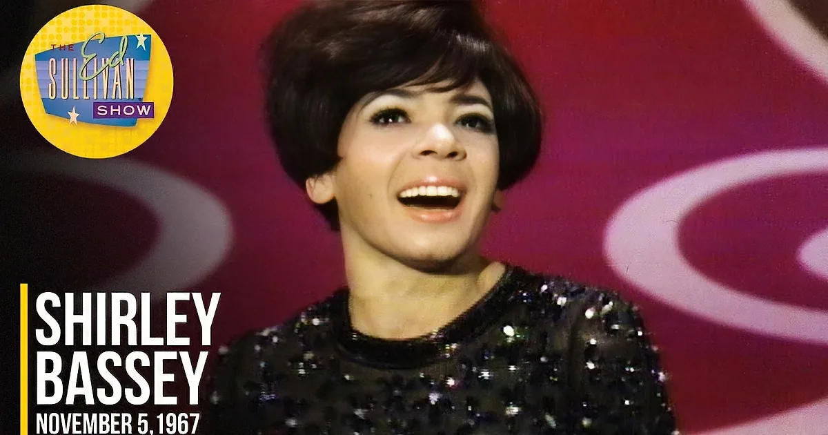 A smiling woman with short, dark hair and a black sequined top stands against a red background with circular patterns.