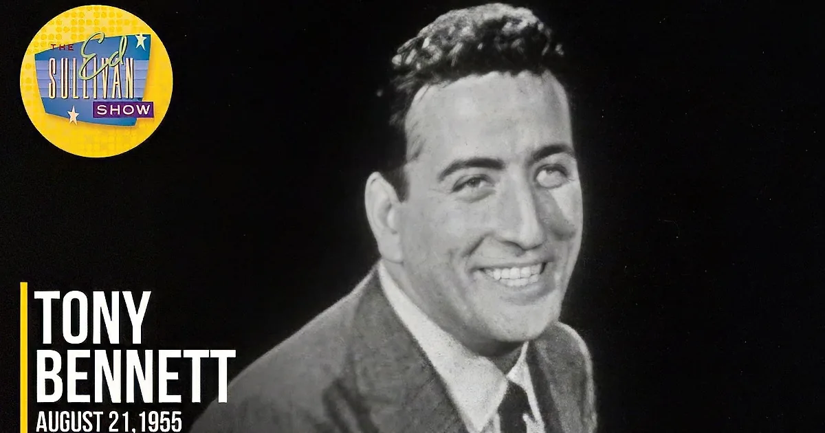 A smiling man in a suit and tie, with a black background and a watermark in the corner.