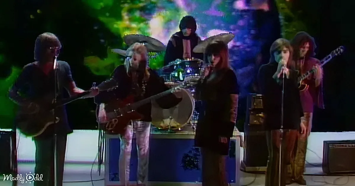 A band performs on stage with colorful lights, featuring a guitarist, drummer, and vocalists.