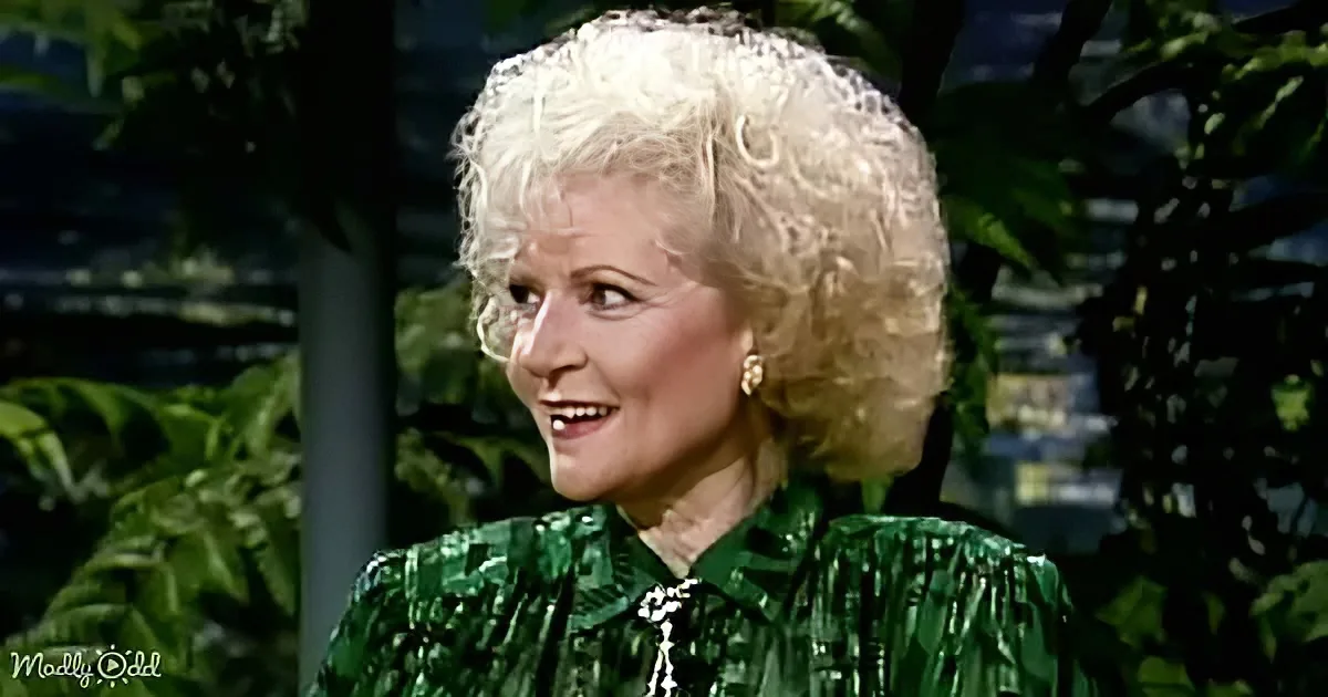 A woman with blonde, curly hair in a green, ruffled top smiles, surrounded by greenery.