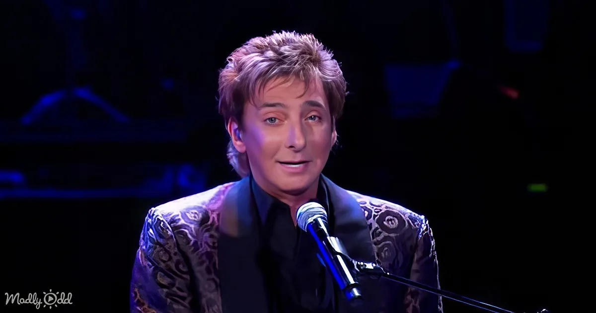 A person in a patterned suit sings into a microphone on stage with a dark background.