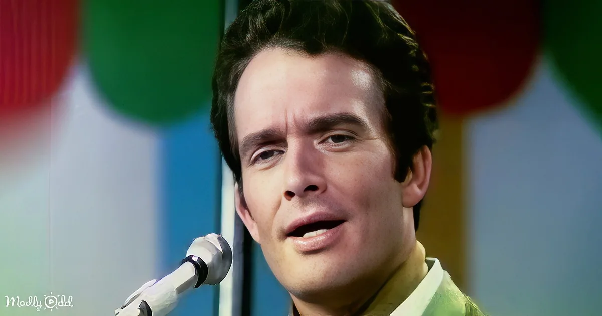 A man with dark hair and a light green shirt sings into a microphone, with colorful balloons in the background.