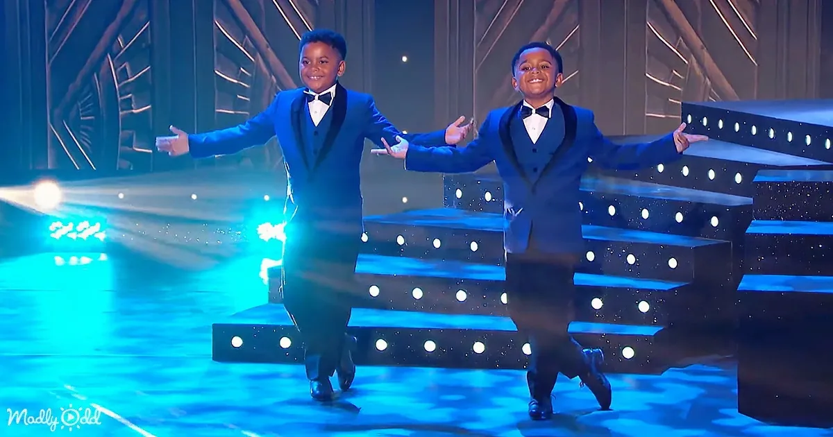 Two boys in blue tuxedos perform on stage with arms outstretched, surrounded by blue lighting and a modern backdrop.