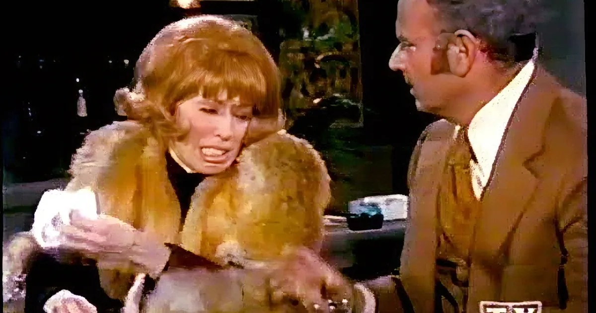 A woman in a fur coat smiles while a man in a suit looks on, both in a dimly lit room.