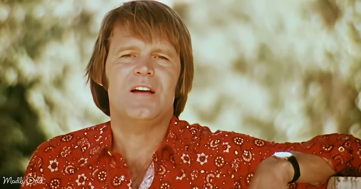 A person with shoulder-length hair, wearing a red patterned shirt, is seen against a blurred background.