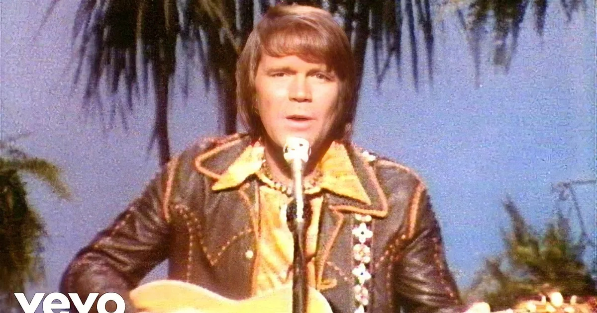 A smiling man with long, wavy brown hair and a microphone, wearing a patterned shirt.
