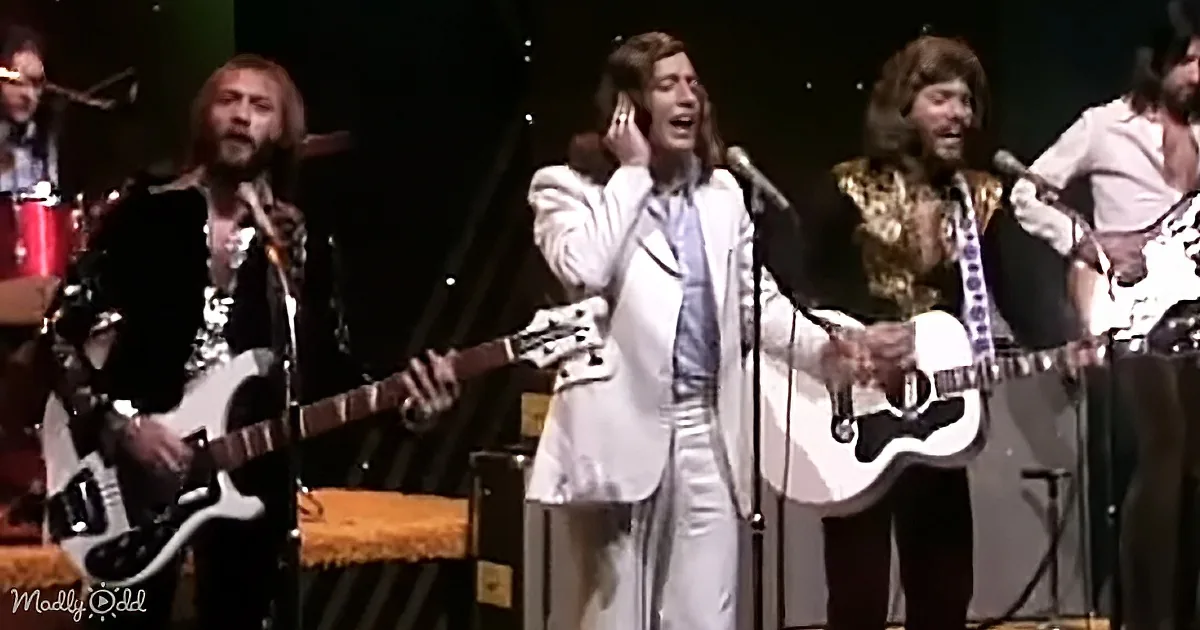 A man in a white suit sings passionately on stage, surrounded by band members playing guitars.