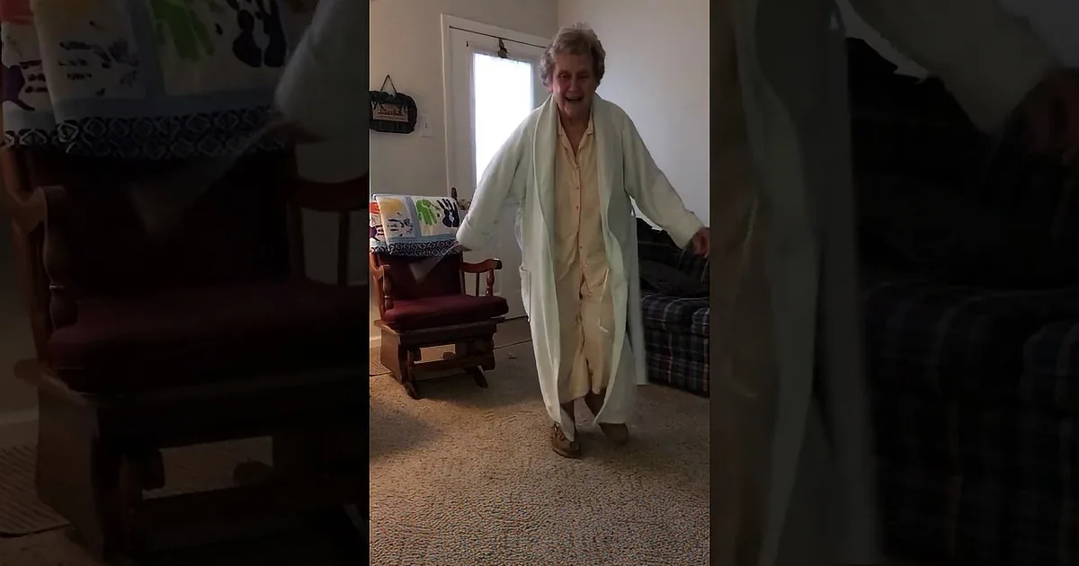 An elderly woman in a white robe dances joyfully in a brightly lit room.