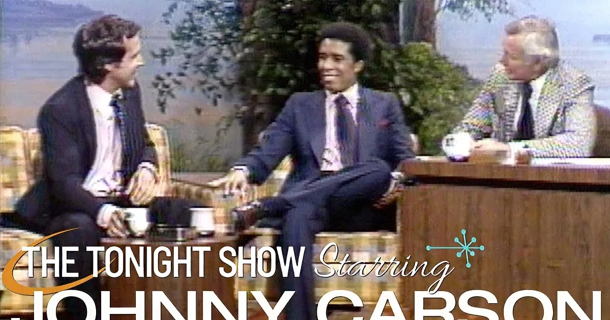 A man in a suit sits between two others on a talk show set, smiling.