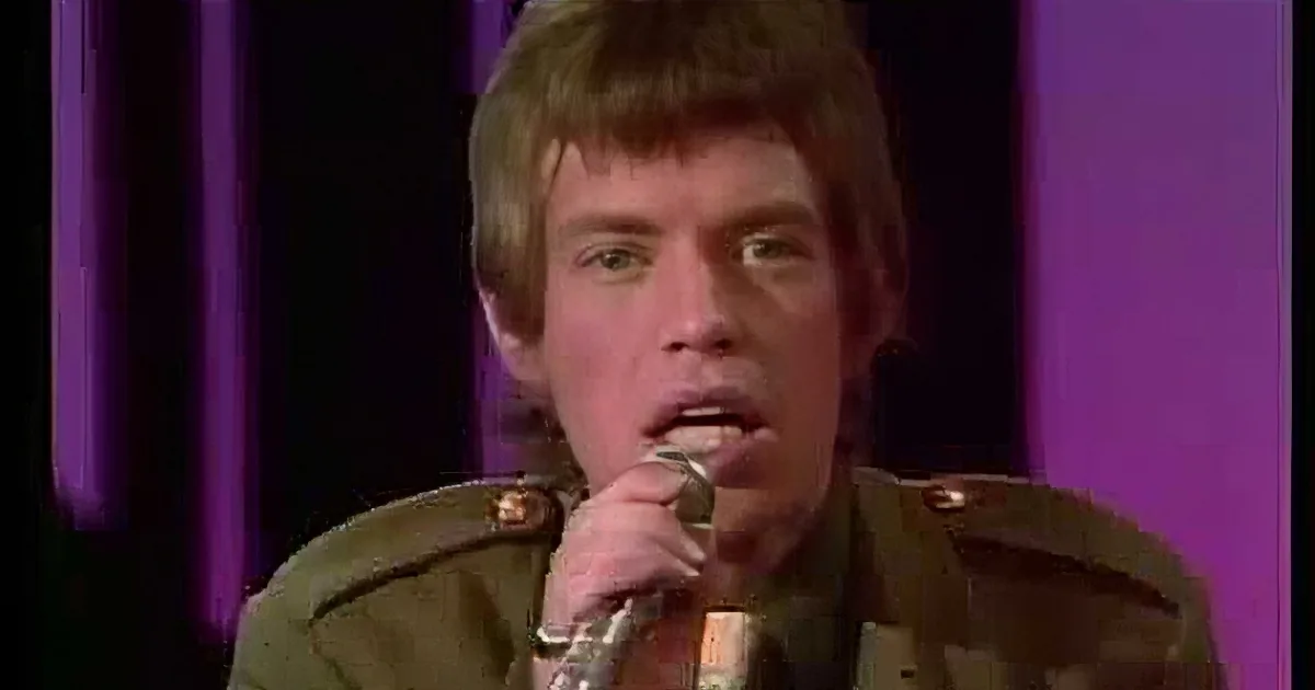 A person with long hair and a ring on their finger holds a microphone, singing in front of a purple backdrop.