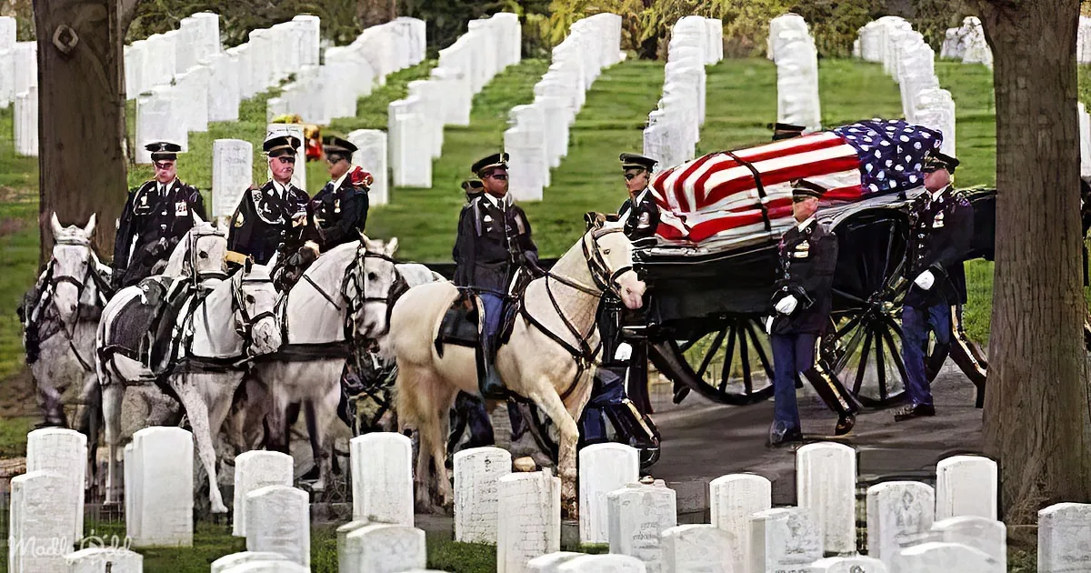 A horse-drawn carriage carrying a casket draped with an American flag leads a procession through a cemetery.