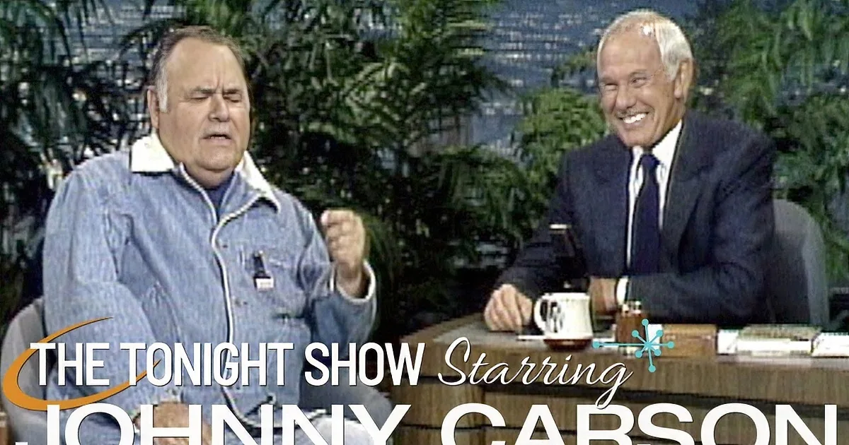 A man in pajamas sits with his fist raised, while another man in a suit smiles across a desk.